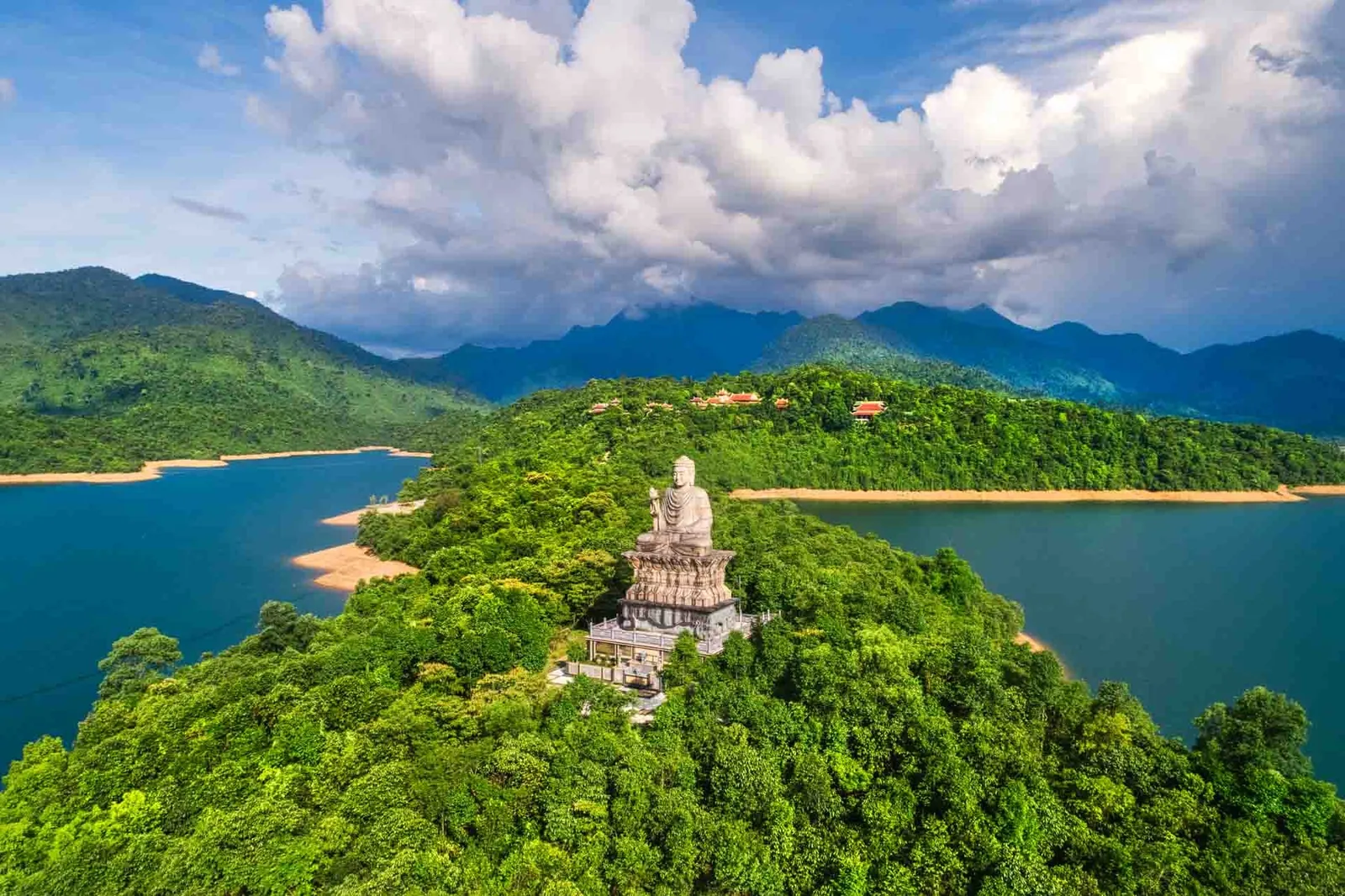 A large white Buddha statue stands atop a forested hill overlooking a reservoir near Bach Ma National Park, Hue.