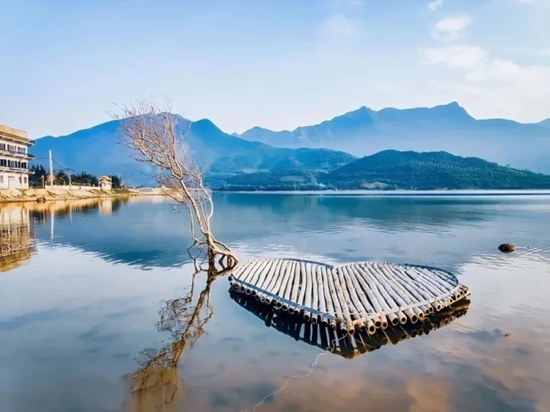 A bamboo raft floats beside a bare tree on the calm waters of Lap An Lagoon with misty mountains behind. A bamboo raft floats beside a bare tree on the calm waters of Lap An Lagoon with misty mountains behind.