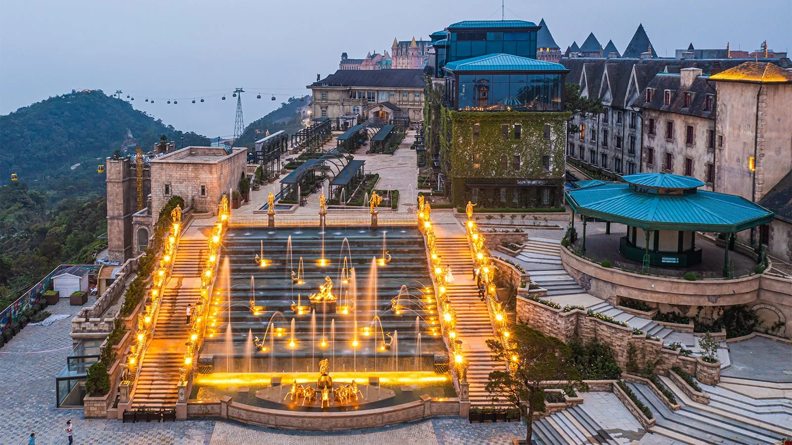 European-style architecture and illuminated fountain staircase at Ba Na Hills at dusk with cable car in the background.