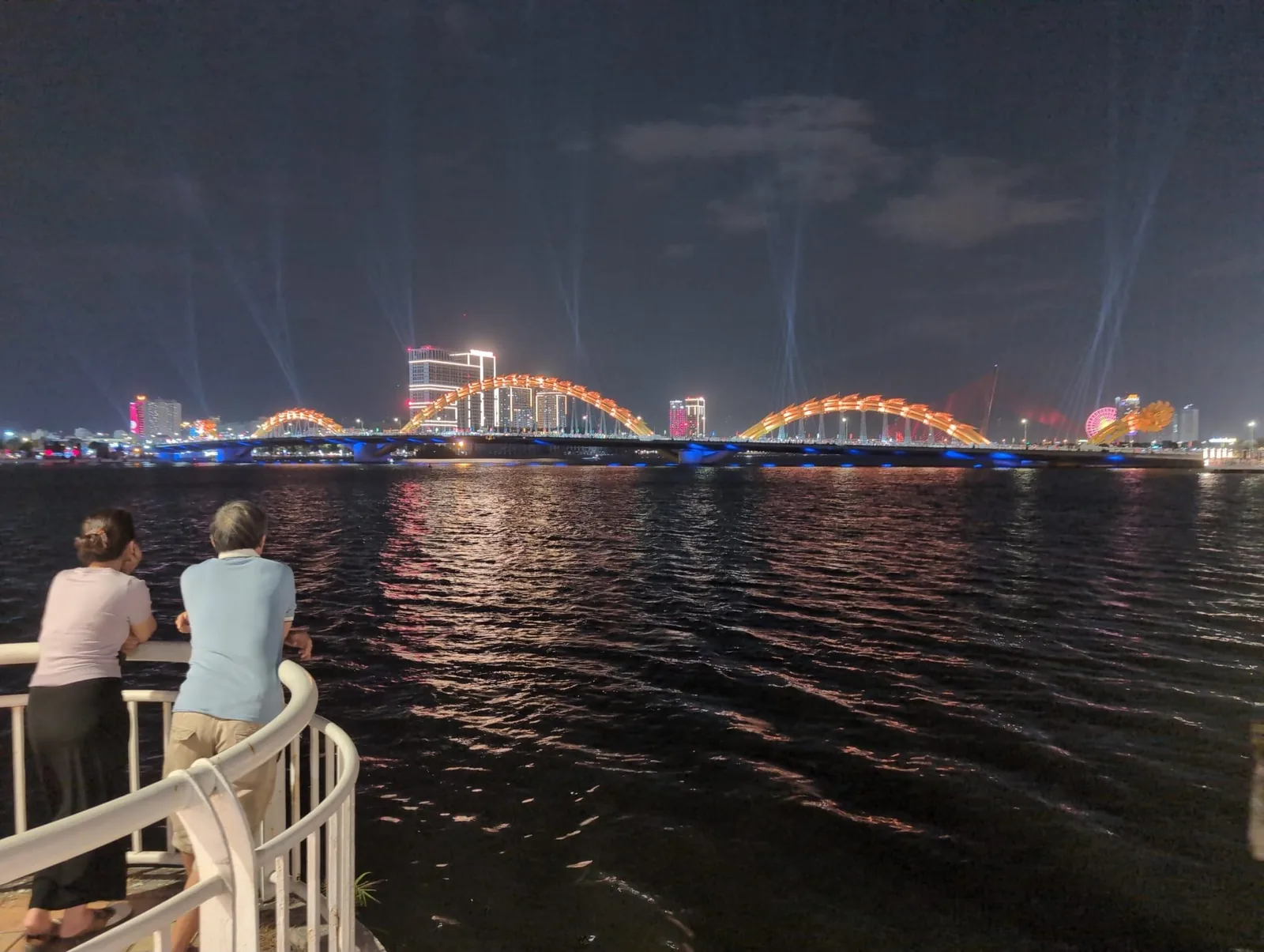 Two people watch the illuminated Dragon Bridge over the Han River in Da Nang at night. Two people watch the illuminated Dragon Bridge over the Han River in Da Nang at night.