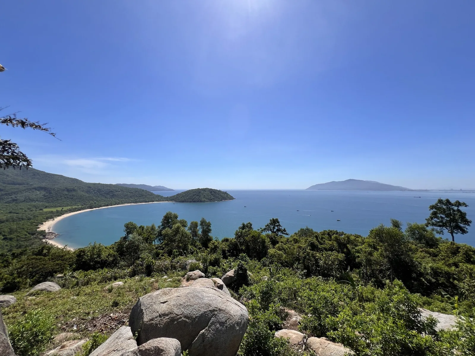 A sweeping coastal view of a curved sandy beach and green hills along the Da Nang shoreline under a clear blue sky. A sweeping coastal view of a curved sandy beach and green hills along the Da Nang shoreline under a clear blue sky.