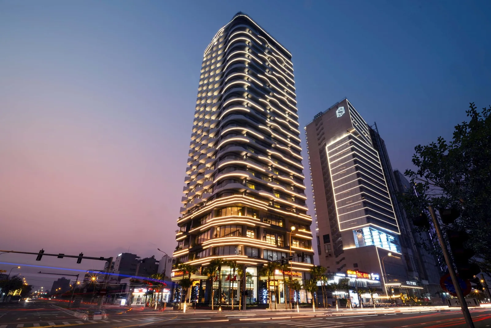 A modern high-rise hotel tower glows with warm lighting at dusk on a Da Nang street.